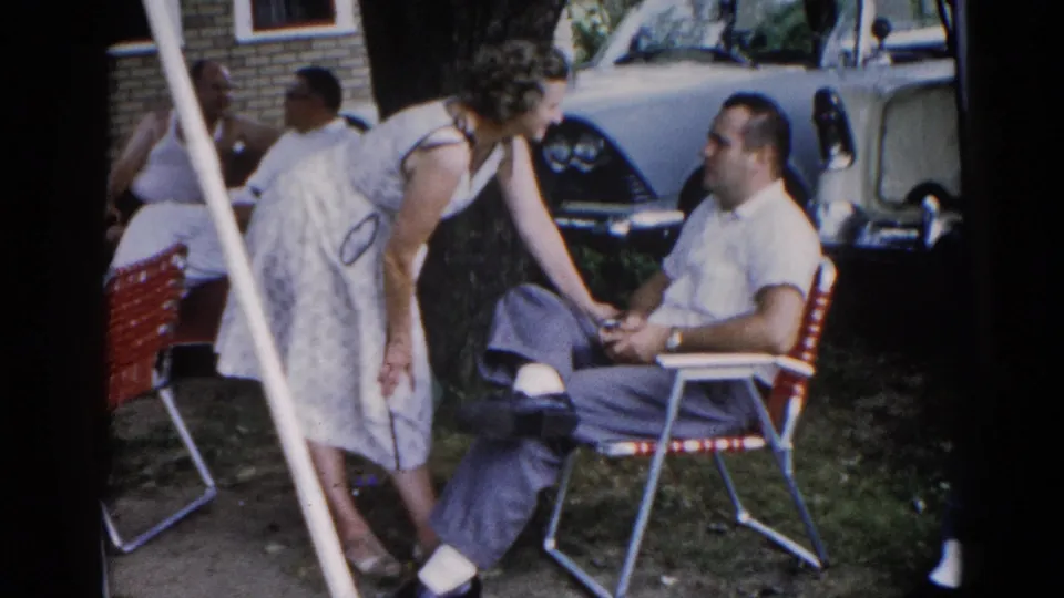 A woman in a white dress speaks to a man seated in a red tubular lawn chair in a suburban backyard; a late-1950s or early-1960s American car is visible behind them, Illinois circa 1962.