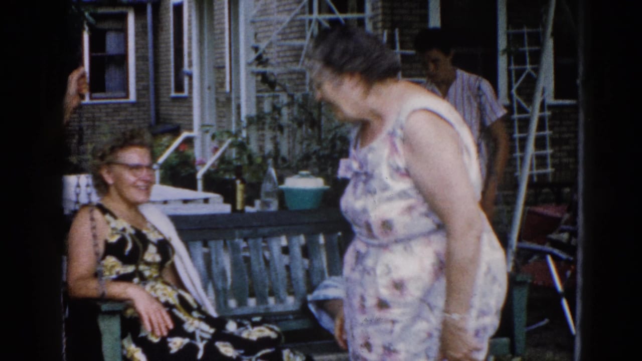 Two women in candid porch conversation — seated woman in dark floral dress with eyeglasses, standing woman in light print dress bending forward; Illinois residential setting, c. 1962.