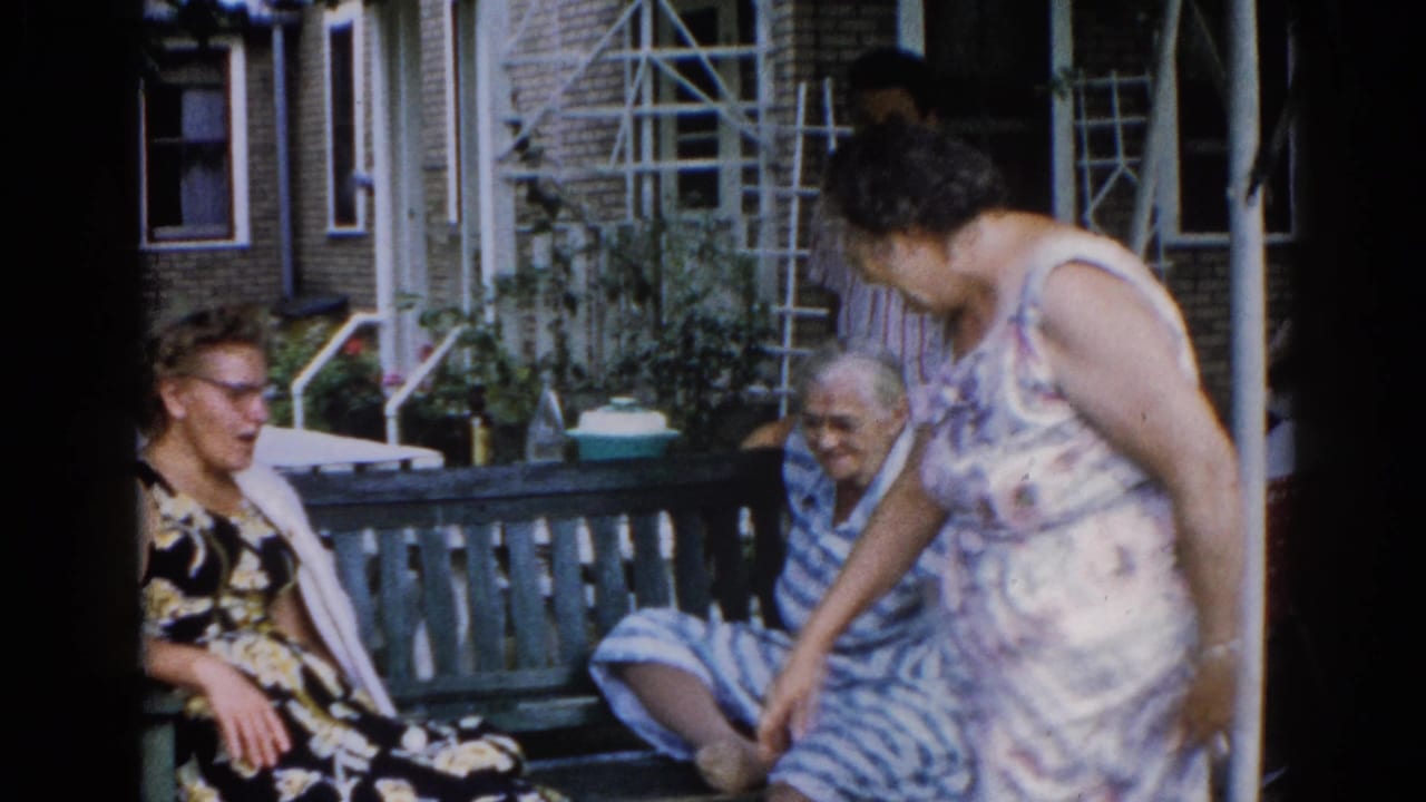 Three women in candid porch conversation, Illinois c. 1962 — standing woman in light print dress leans toward older woman in striped garment; man in striped shirt and brick facade behind them.