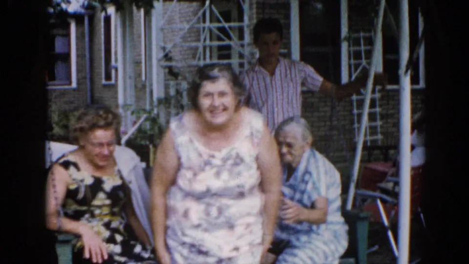Close-medium view of two smiling women on a residential porch in Illinois, c. 1962; older woman at right edge and a man in striped shirt visible in background.