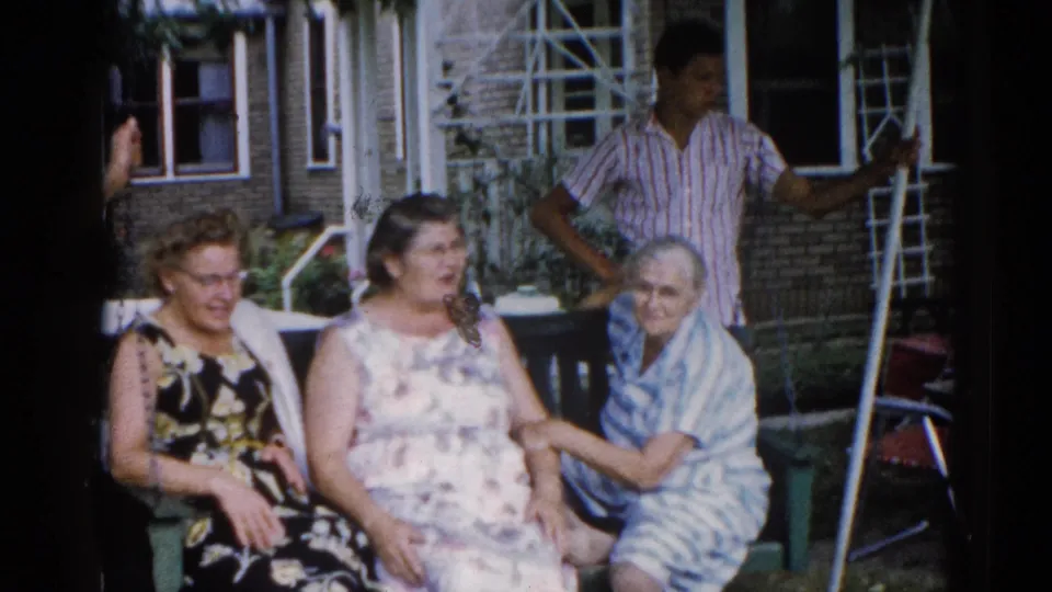 Three women in animated conversation and laughter on a residential porch, Illinois c. 1962; man in striped shirt and brick building facade visible in background.