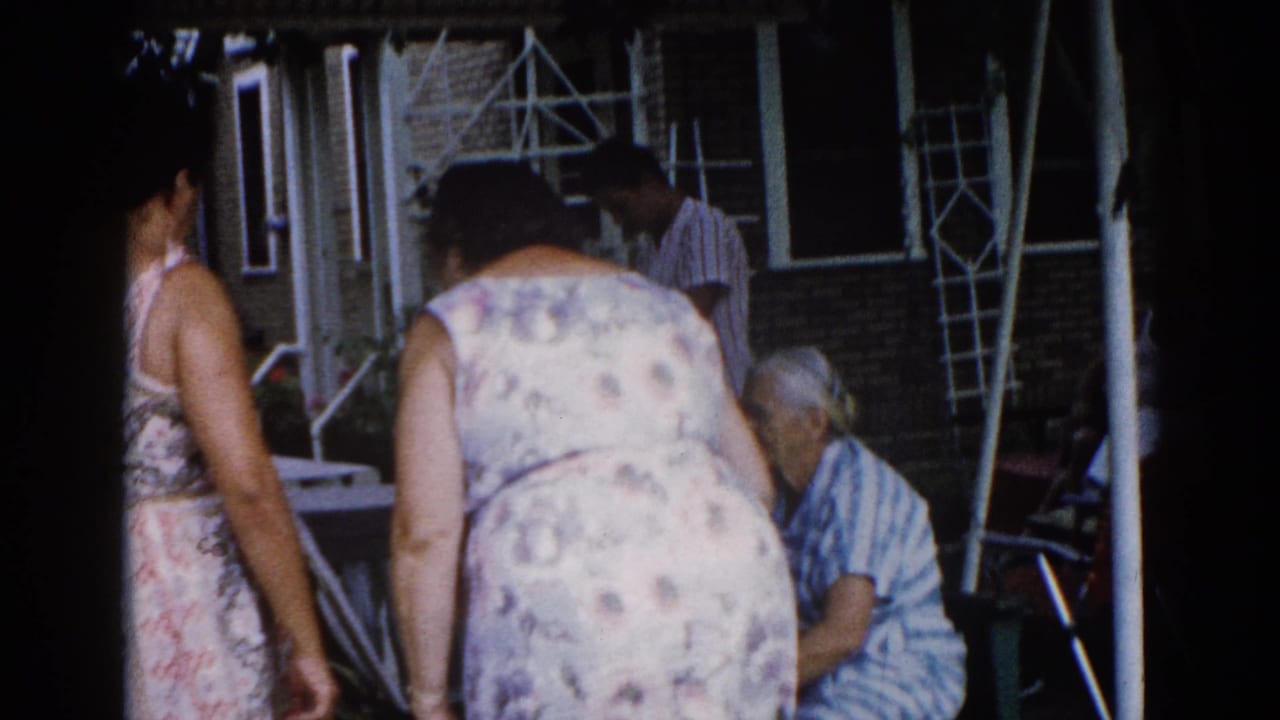 Group of women outdoors beside a brick house; central figure in floral print dress stands with back to camera.