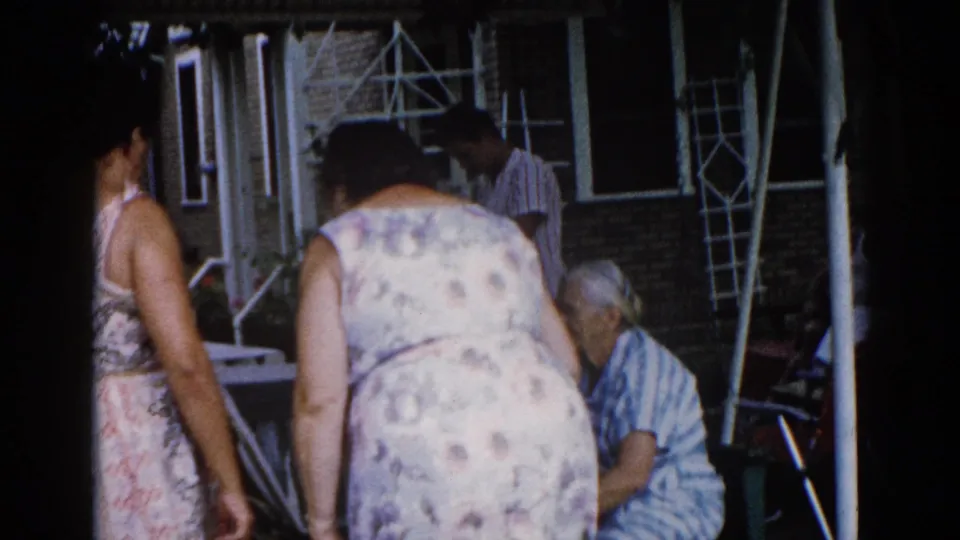 Women gathered outside a brick residential building in what appears to be Illinois, circa 1962; 8mm home movie, central figure in floral dress facing away.