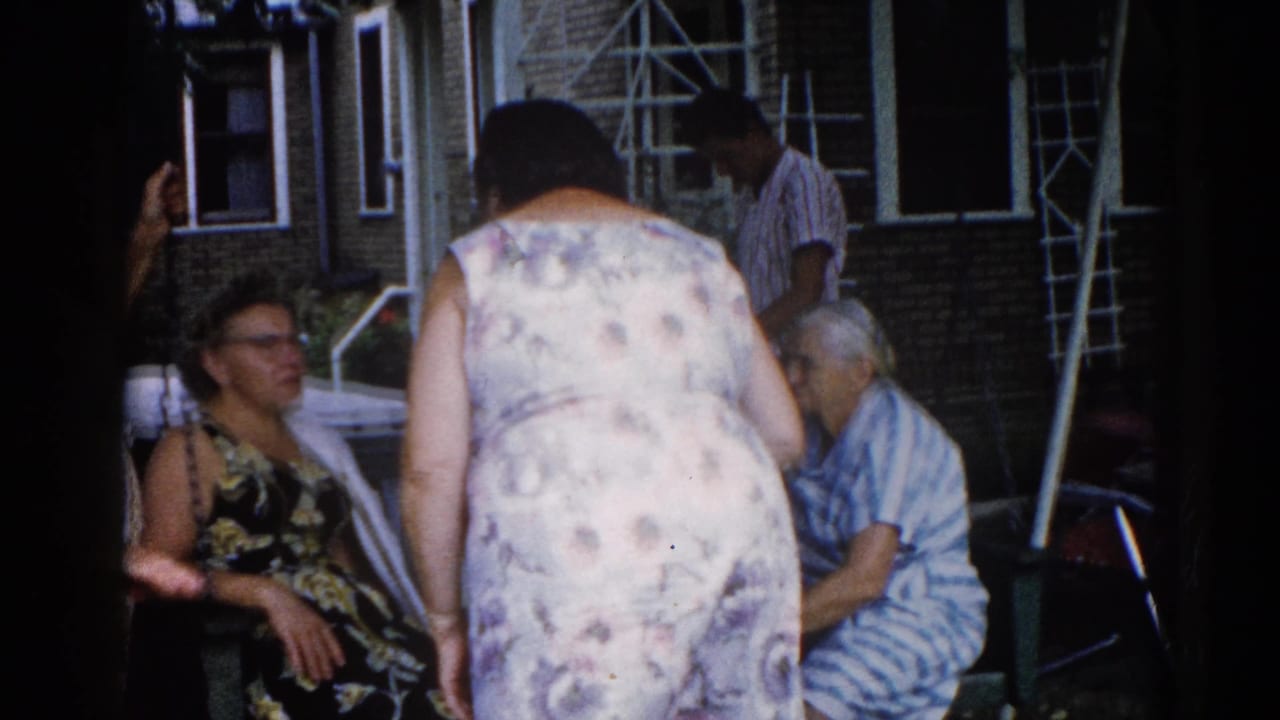 Women socializing outside a brick house; seated woman in black floral dress with glasses visible at left, central figure in floral dress standing.
