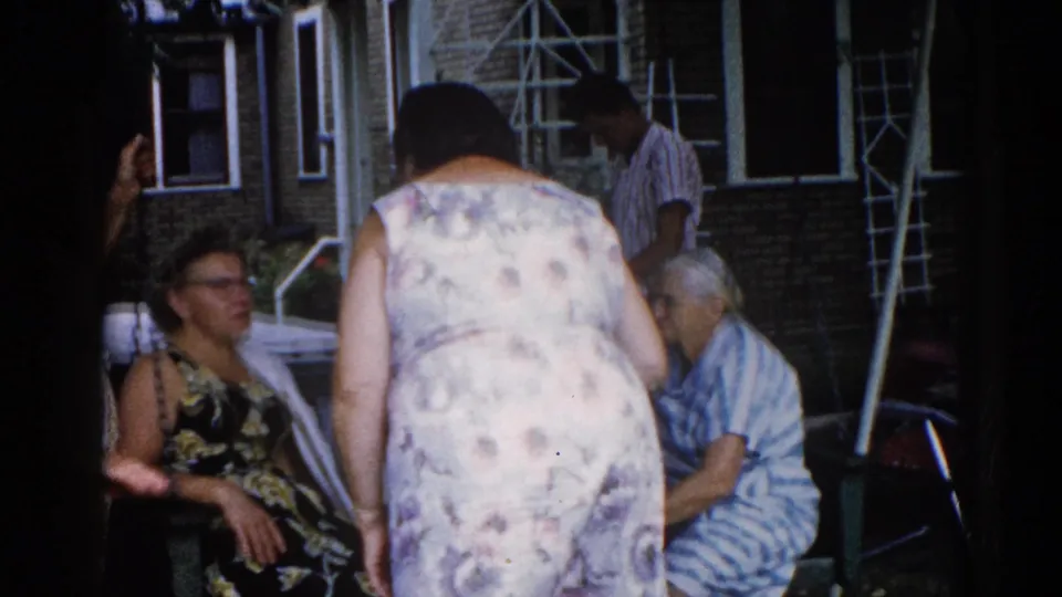 Candid group of women outside a brick house, circa 1962 Illinois; woman in glasses seated at left, central figure in floral dress standing with back to camera.