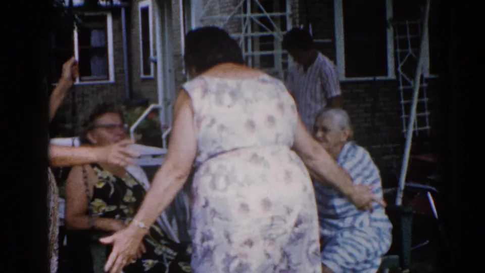 Candid 8mm home-movie frame of women in conversation outside a brick house, circa 1962; central figure bending toward seated companions in summer dresses.
