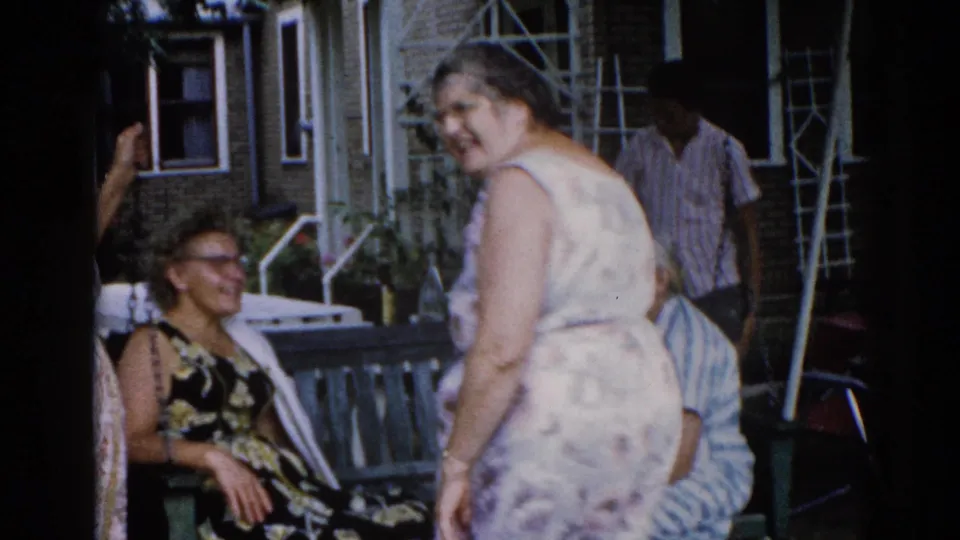 Woman in 1960s floral dress turns to smile at the camera in an unguarded home-movie moment; seated woman with glasses and man in striped shirt visible behind, brick house exterior, circa 1962 Illinois.