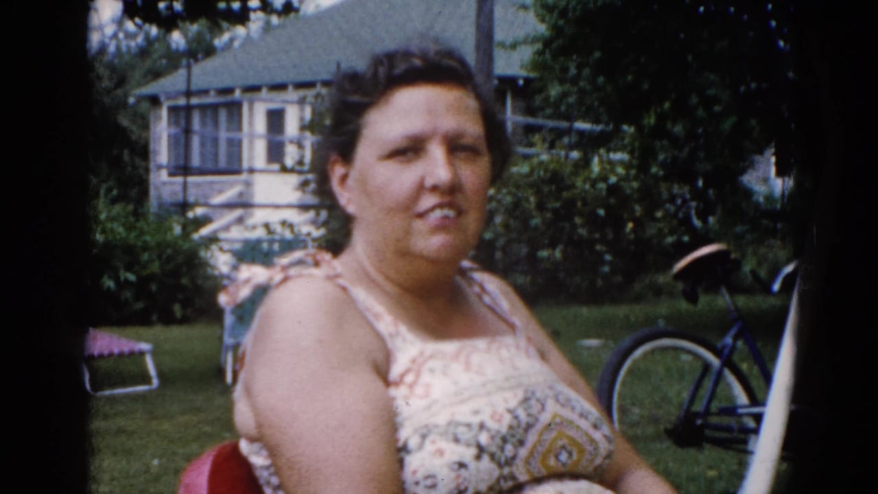 Woman in floral sundress looks toward camera in a suburban backyard, consistent with early-1960s Illinois; bicycle, trees, and neighboring house behind her.