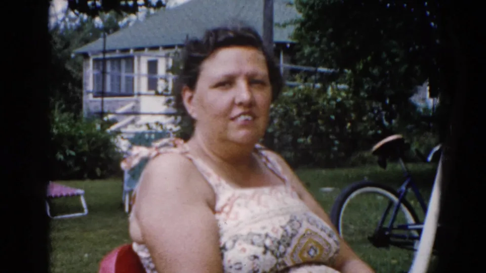 Woman in floral sundress looks toward camera in a suburban backyard, consistent with early-1960s Illinois; bicycle, trees, and neighboring house behind her.
