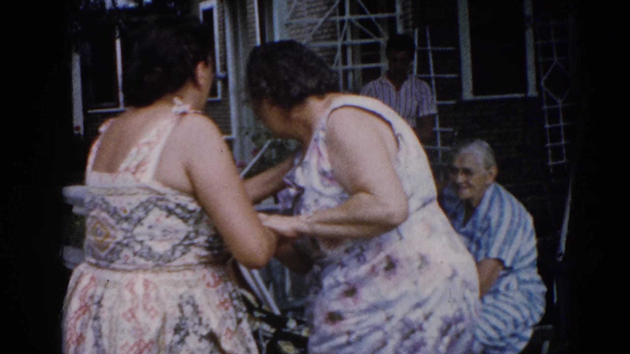 Two women in floral sundresses share an intimate moment in backyard, circa 1962 Illinois; older man in striped shirt watches from a chair near brick house wall.