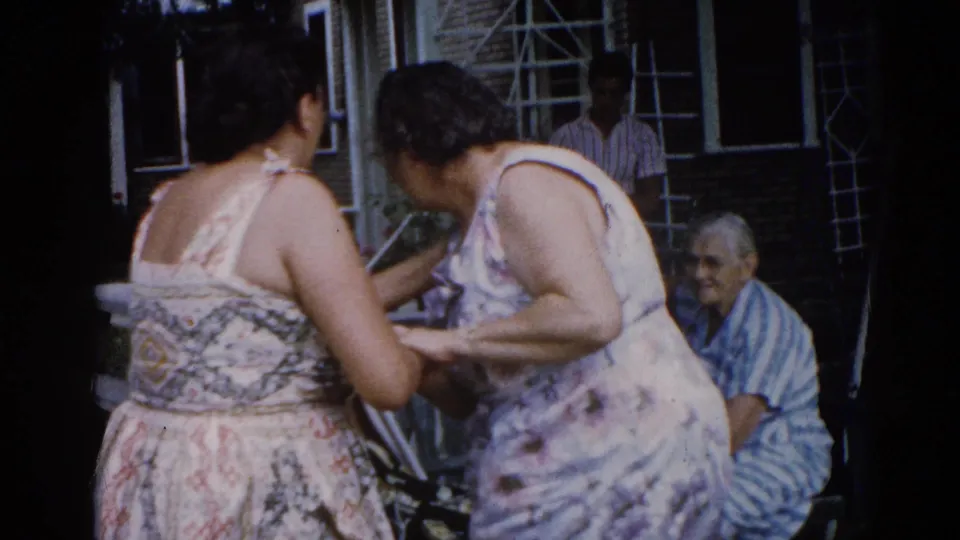 Two women in floral sundresses share an intimate moment in backyard, circa 1962 Illinois; older man in striped shirt watches from a chair near brick house wall.