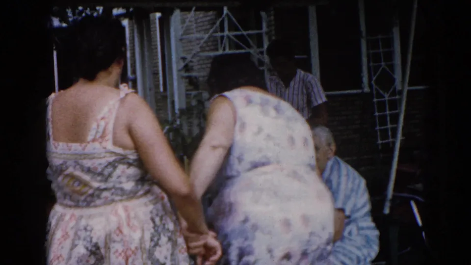 Two women in floral dresses stand very close together, shot from behind in a backyard; older man in striped shirt seated at right, brick structure behind them, circa 1962.
