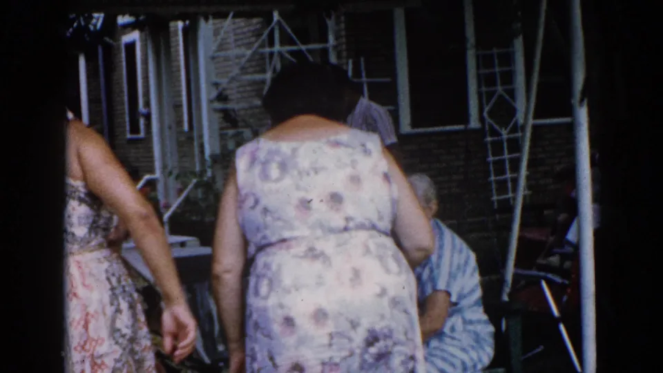 Two women in sundresses stand close together near brick house, shot from behind; ladder visible at right, older man seated nearby, circa 1962 Illinois.