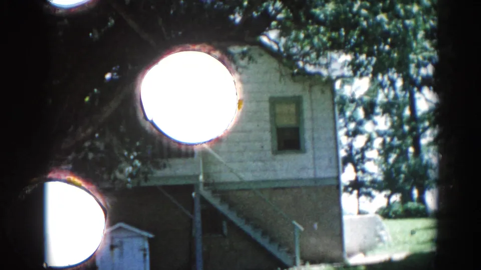 Establishing wide shot of an Illinois backyard circa 1962: frame house with green-gray roof, rear stairs, shed, and mature deciduous trees.