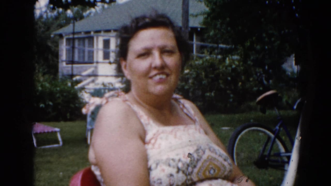 A woman in a patterned sundress sits in a red metal lawn chair smiling at the camera; Illinois suburban backyard circa 1962, with bicycle and frame house behind.
