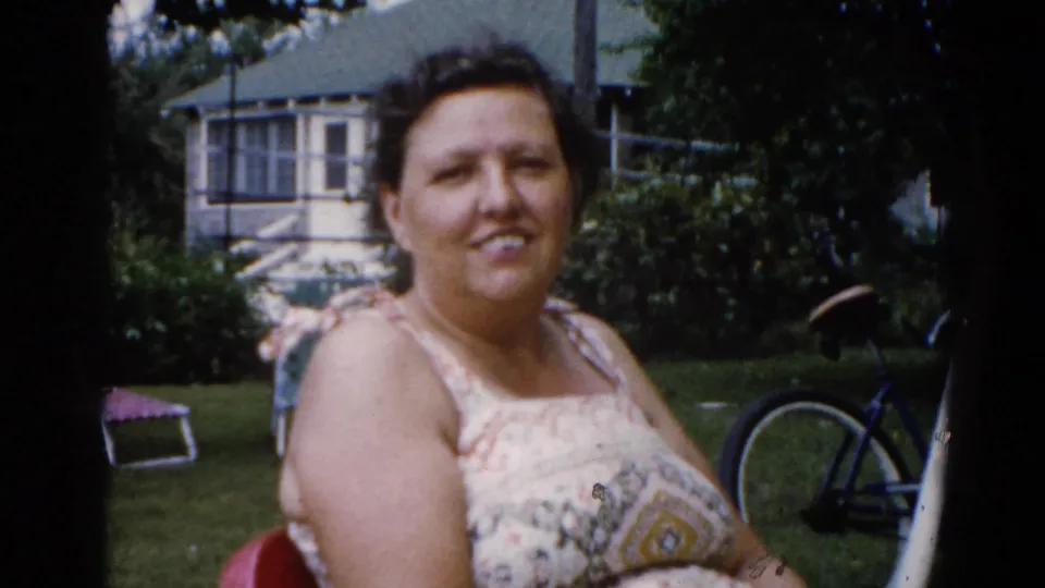 A woman in a patterned sundress sits in a red metal lawn chair smiling at the camera; Illinois suburban backyard circa 1962, with bicycle and frame house behind.