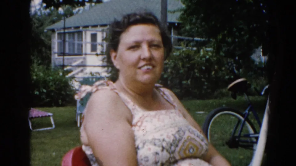 Woman in patterned sleeveless sundress seated in a red lawn chair, facing the camera; Illinois backyard circa 1962 with frame house, screened addition, and bicycle behind.