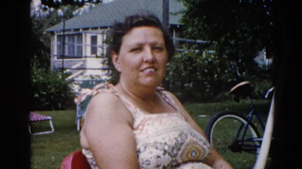 Near-identical composition to previous frame — woman in patterned sundress in red metal lawn chair, smiling at camera; Illinois backyard circa 1962.