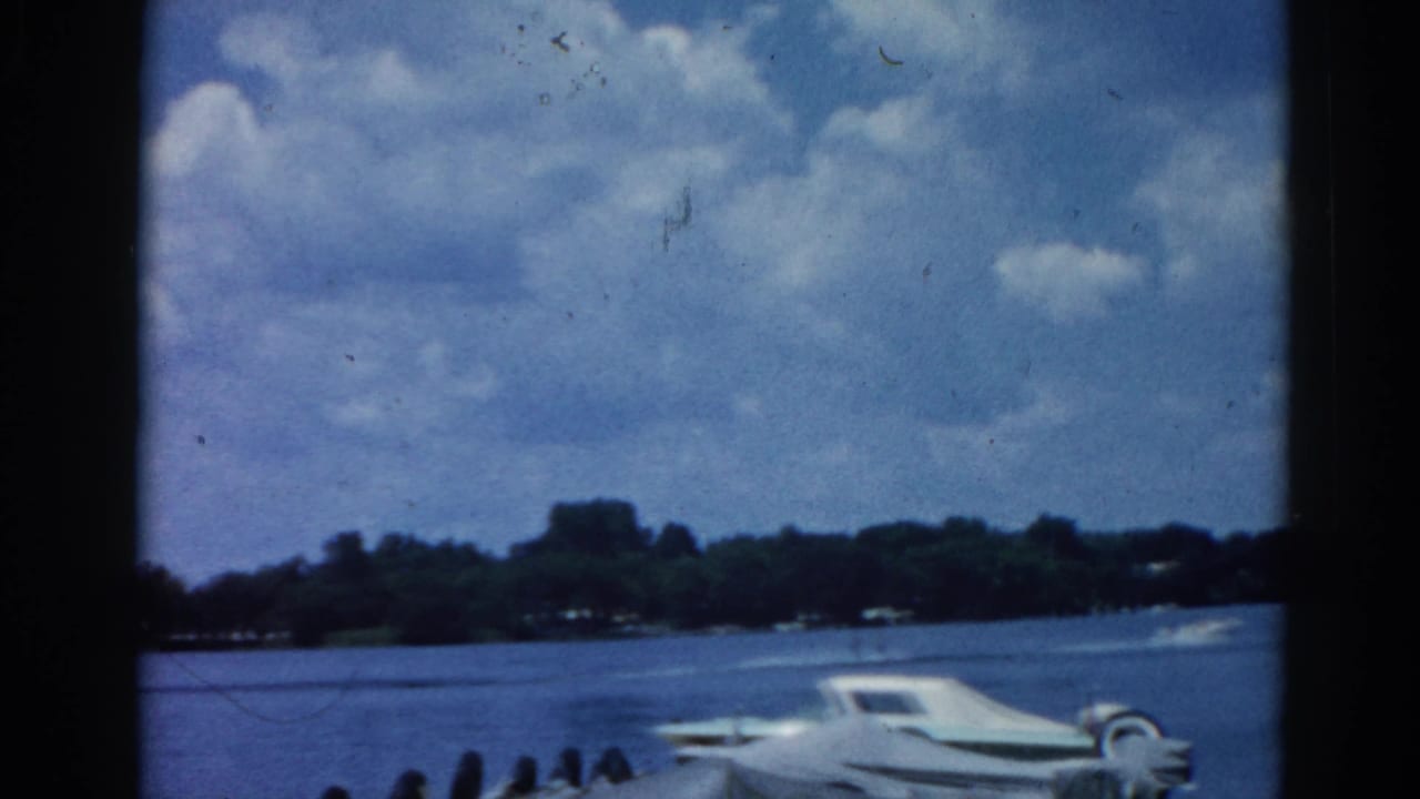 White motorboat moves across a calm lake under partly cloudy sky; Grain Belt Beer sign text is partially visible at the lower frame edge, tree-lined far shore in the distance.