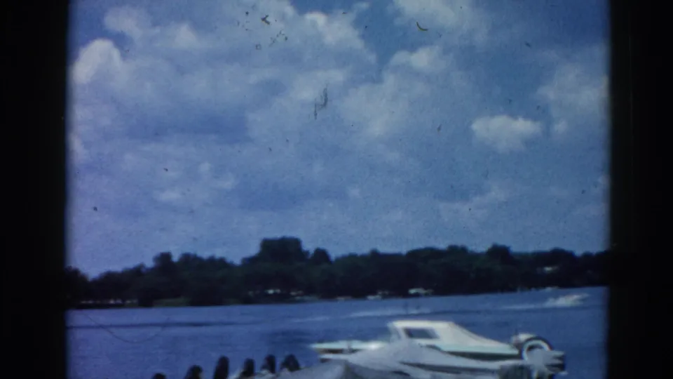 8mm home movie frame circa 1962, Illinois: white motorboat on calm lake, cumulus clouds, tree-lined shore, with Grain Belt Beer Premium sign partially cropped at the lower edge.