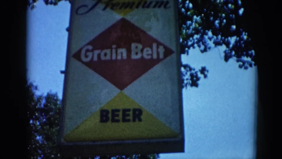 Grain Belt Beer Premium sign, slightly wider framing circa 1962, Illinois: full diamond logo readable with surrounding tree canopy and open sky visible around the sign.