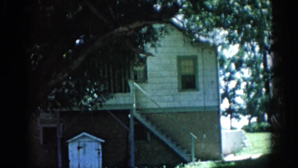 Modest two-story residential building circa 1962, Illinois: exterior wooden staircase, white-painted upper siding, brick lower section, small shed or outbuilding, mature deciduous trees.