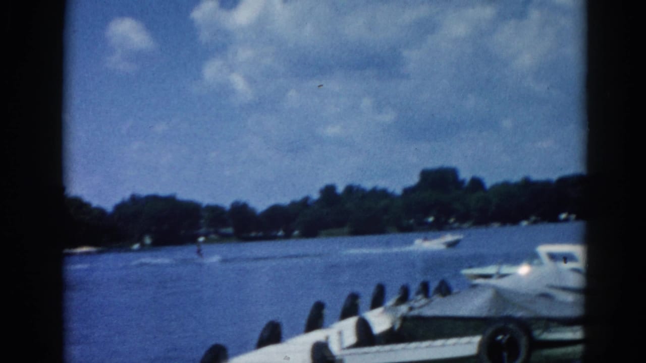 Lake scene with white motorboat moored at dock right, open water, wooded far shore, partly cloudy summer sky.