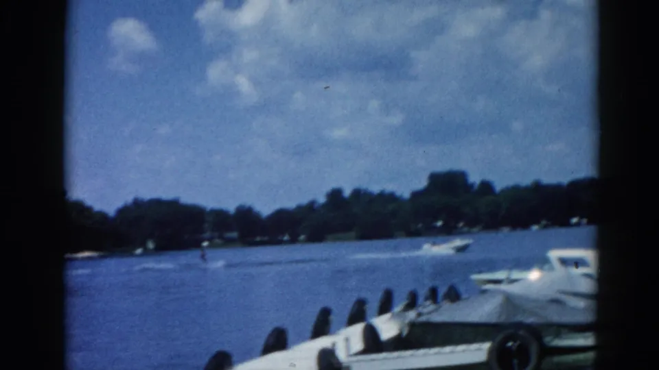 8mm home movie frame circa 1962, Illinois lake with white motorboat moored at dock, tree-lined far shore and cumulus clouds visible across open water.