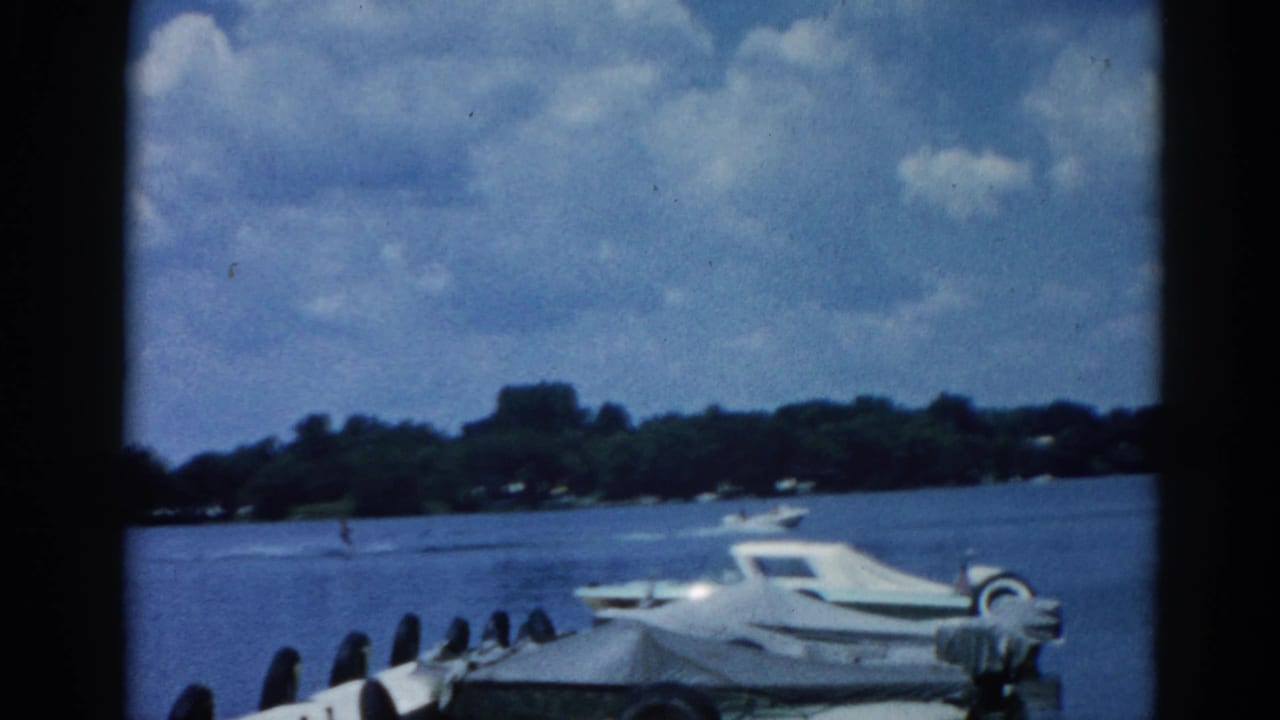 8mm home movie frame circa 1962, Illinois lake with white cabin cruiser moored at dock, summer cumulus clouds dominating the upper frame above a tree-lined shore.