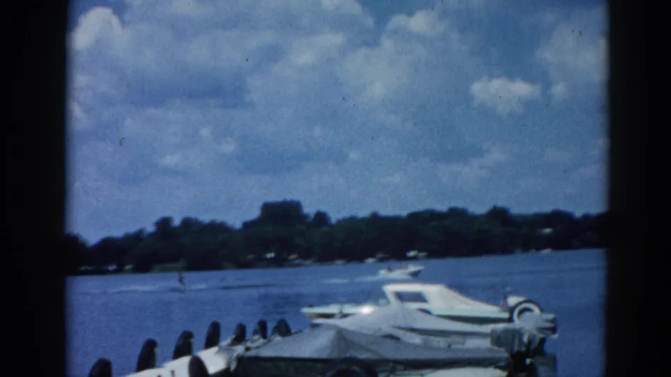 8mm home movie frame circa 1962, Illinois lake with white cabin cruiser moored at dock, summer cumulus clouds dominating the upper frame above a tree-lined shore.