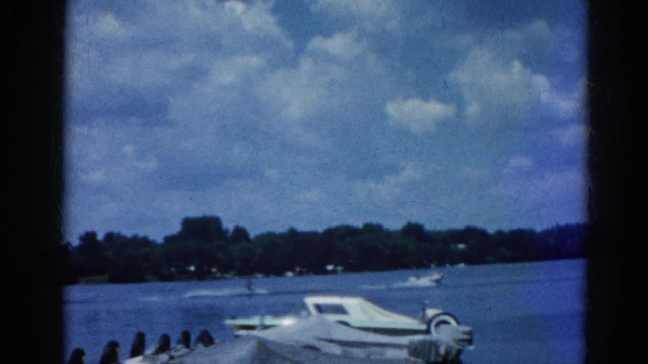 8mm home movie frame circa 1962, Illinois lake with white motorboat at dock, tree-lined shore and cumulus clouds in background across open water.