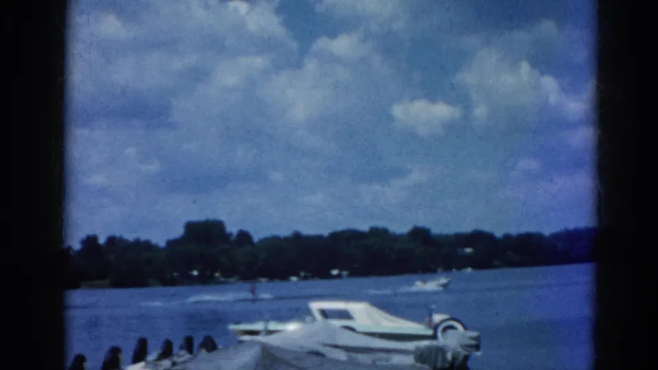 8mm home movie frame circa 1962, Illinois lake with white motorboat at dock, tree-lined shore and cumulus clouds in background across open water.