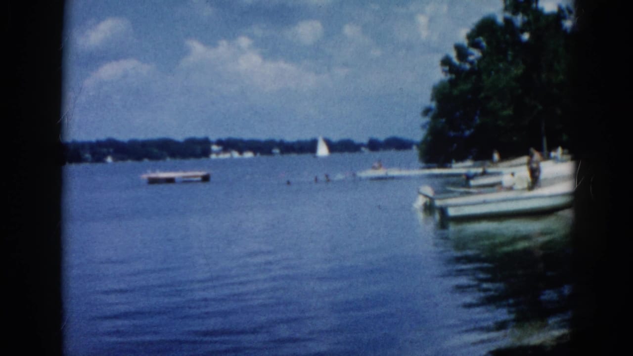 8mm home movie circa 1962, Illinois: calm lake, small motorboats at a dock, white sailboat in the middle distance, partly cloudy sky, and dense green treeline.