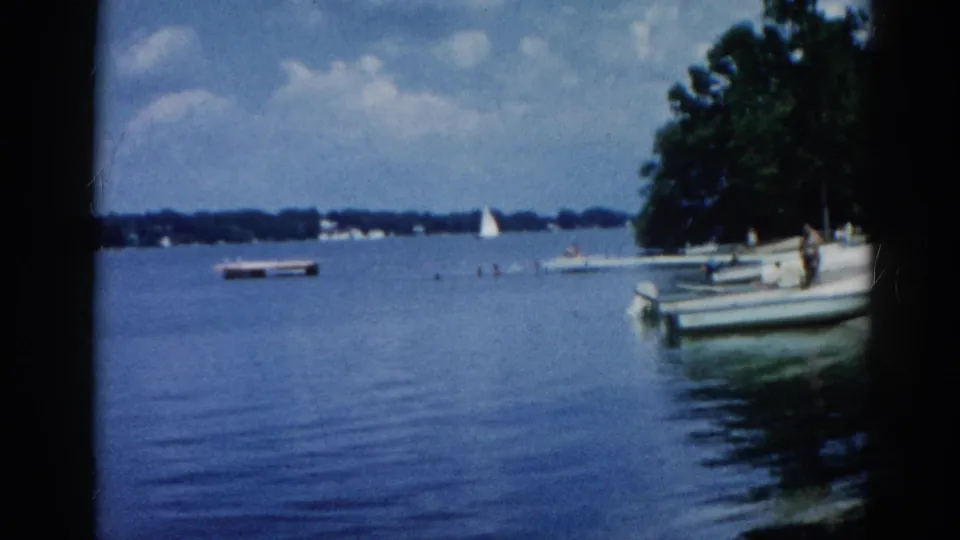8mm home movie circa 1962, Illinois: calm lake, small motorboats at a dock, white sailboat in the middle distance, partly cloudy sky, and dense green treeline.