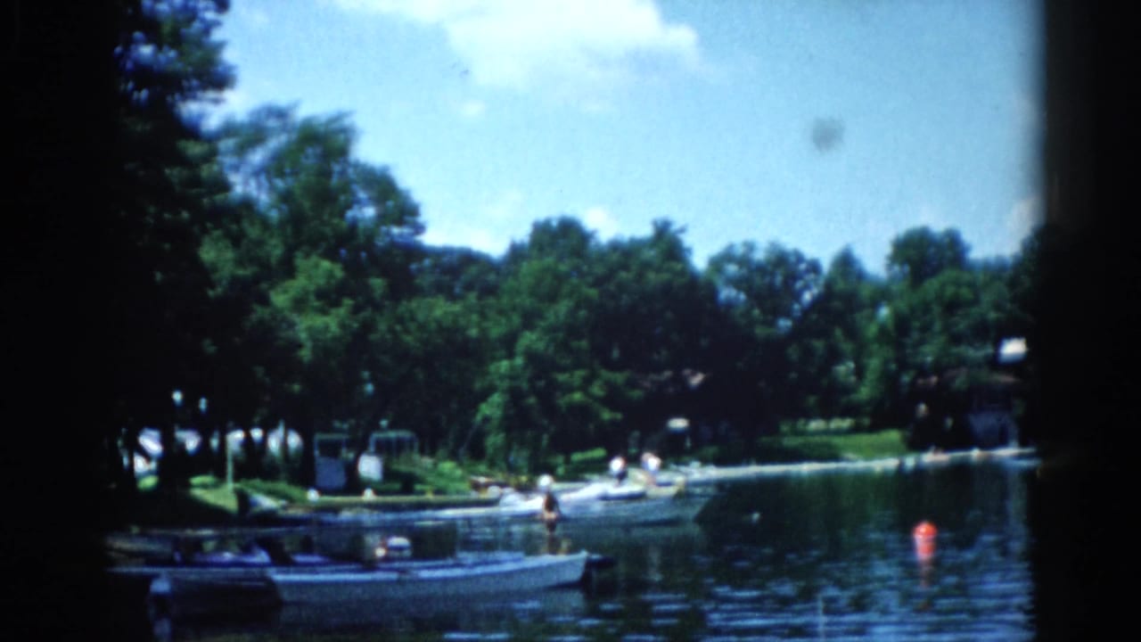 Boat dock with several small motorboats moored, people visible nearby, green trees and houses behind, on 8mm home movie film.