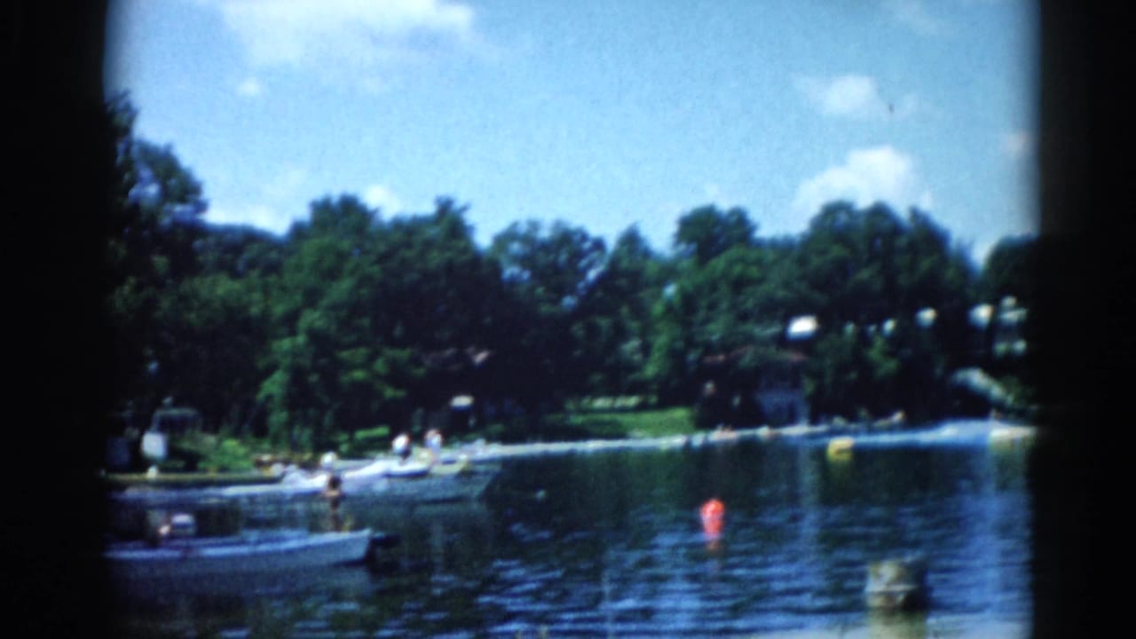 8mm home movie circa 1962, Illinois: open lake with small motorboats near a wooded shoreline, figures at the water's edge, houses among trees, and a red buoy.