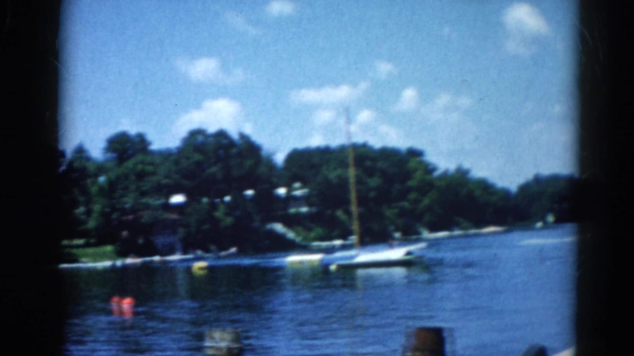 8mm home movie circa 1962, Illinois: lake scene with a sailboat at right, dense tree-lined shore with houses, small motorboats near the bank, and cumulus clouds in a blue sky.