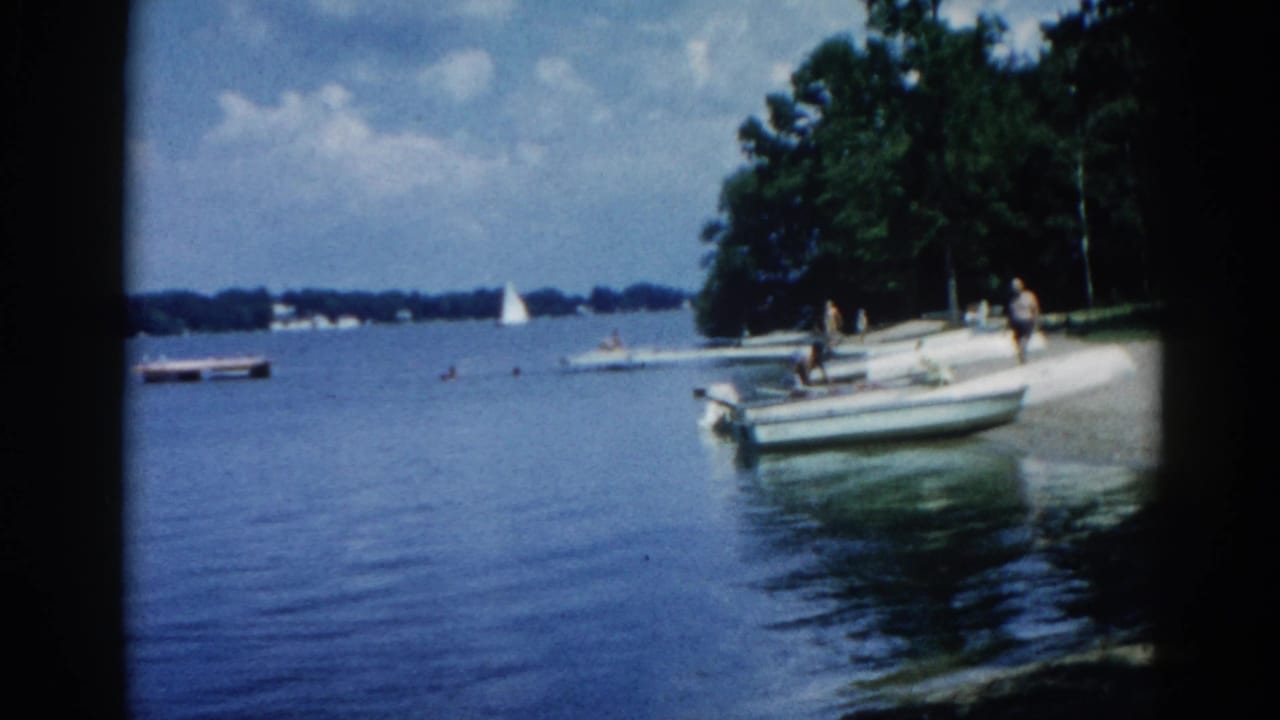 Adults near a motorboat at a lake shore, with sailboats and trees visible in the background.