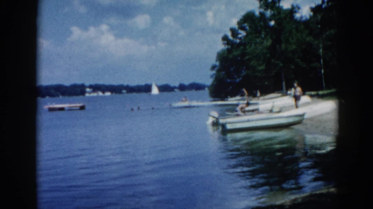 Adults standing near a moored motorboat on a calm lake, with trees and a sailboat in the background.