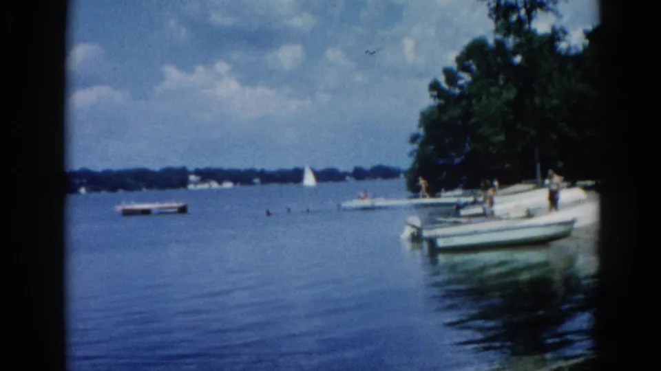 Wide lakeside view with a motorboat and adult figures at the water's edge, Illinois, circa 1962, sailboat visible in distance.