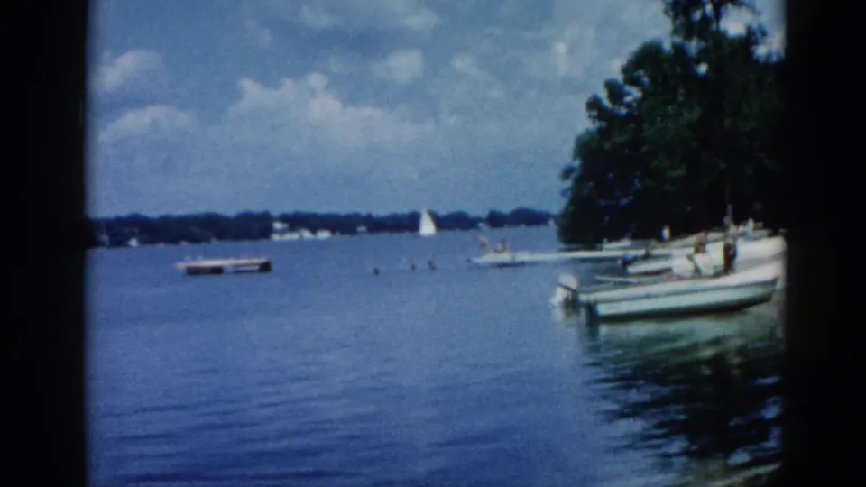Adults near a motorboat at an Illinois lake shore, circa 1962, with distant sailboats and a tree line on the far bank.