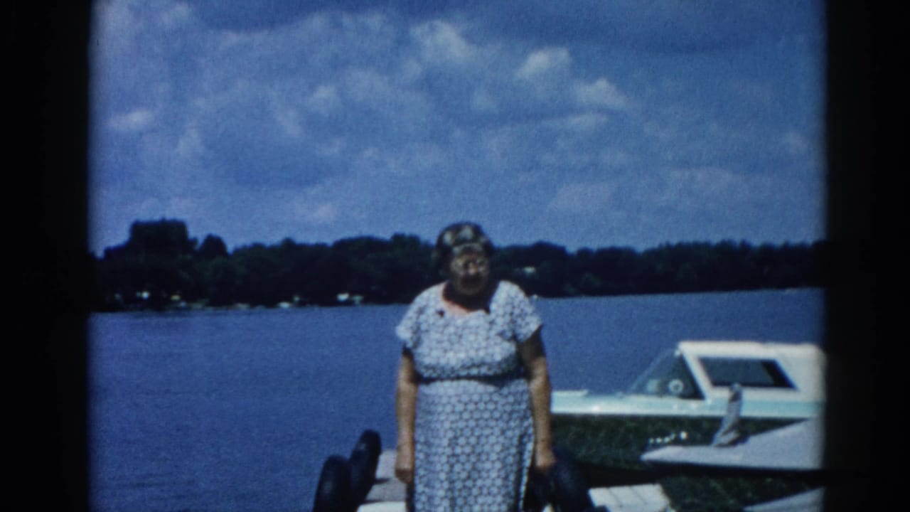 Older woman in a patterned housedress posed on a wooden dock beside a white motorboat at an Illinois lake, circa 1962; 8mm color home movie.