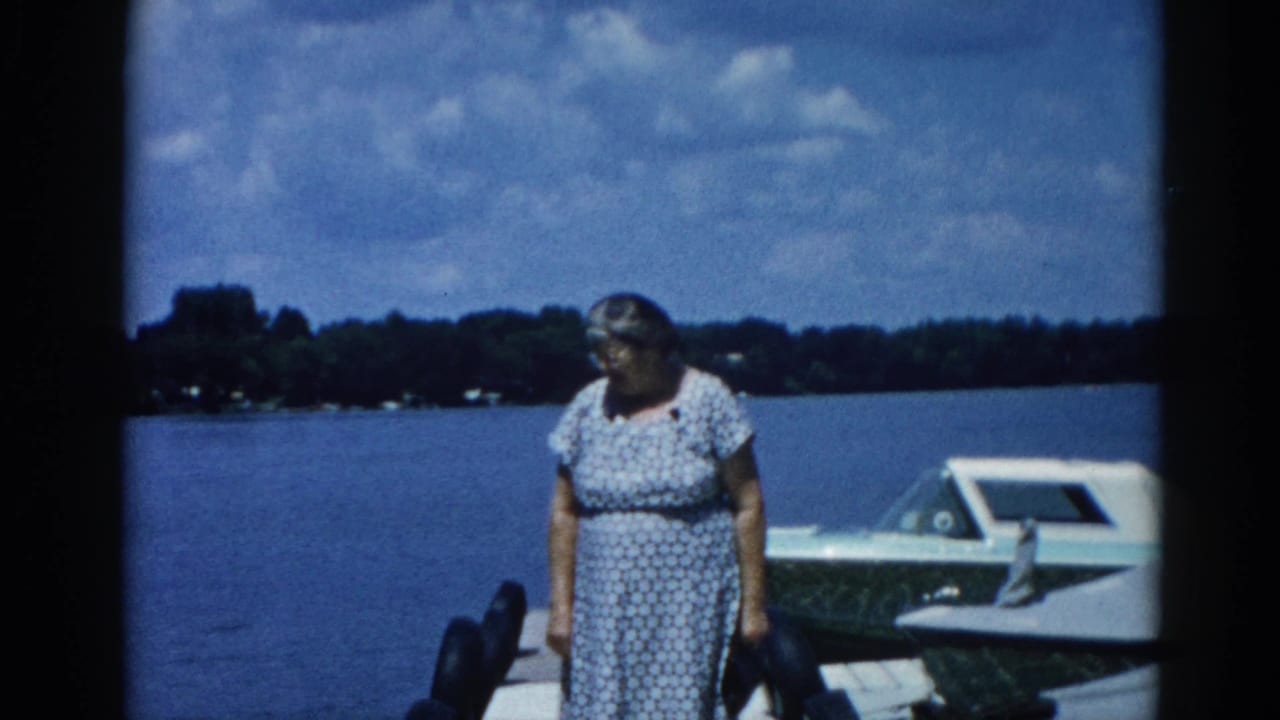 Older woman with glasses standing on a dock beside a white motorboat at an Illinois lake, circa 1962; 8mm home movie color frame.