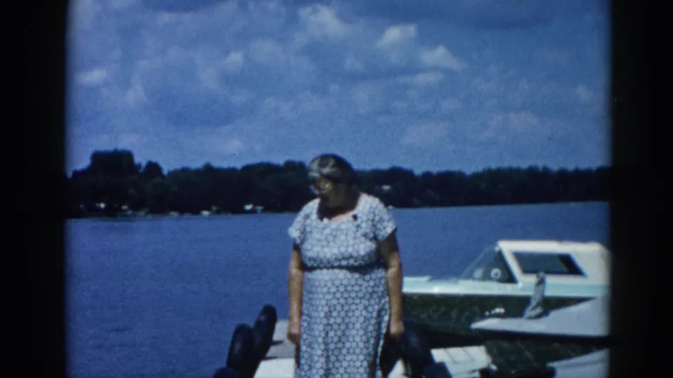 Older woman with glasses standing on a dock beside a white motorboat at an Illinois lake, circa 1962; 8mm home movie color frame.