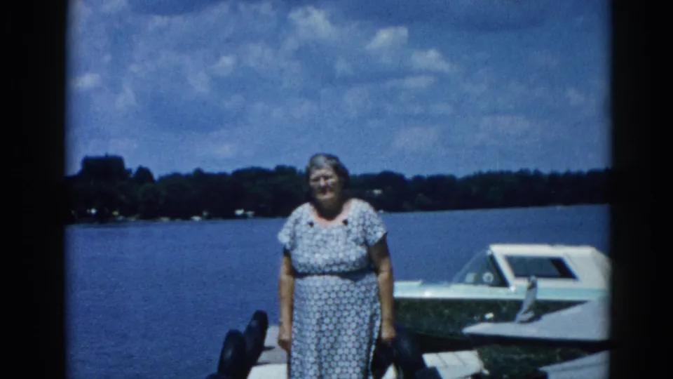 Older woman smiling on a wooden dock beside a moored white motorboat at an Illinois lake, circa 1962; 8mm color home movie.