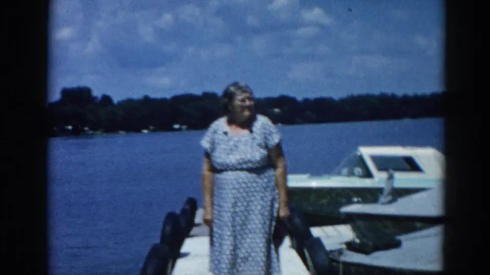 Older woman standing on a dock at an Illinois lake beside a white motorboat, circa 1962; 8mm home movie color footage.