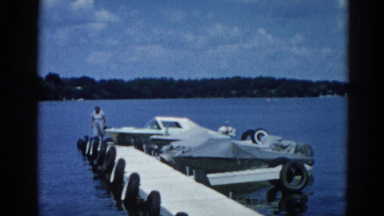 Wide shot of a wooden dock with a white motorboat and canvas-covered boats moored alongside; a woman stands at the far end.