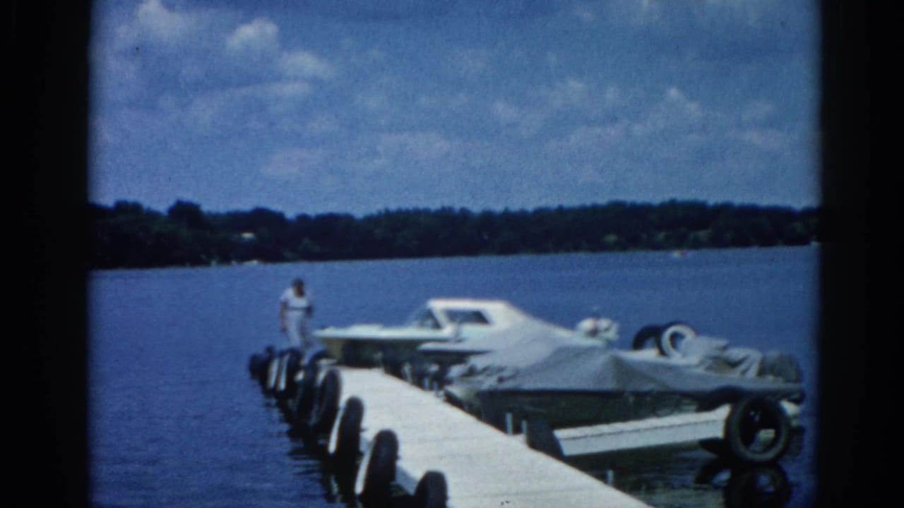 8mm shot of an Illinois lake dock circa 1962; woman standing near covered boats with tire bumpers, white motorboat, and tree-lined shore in background.