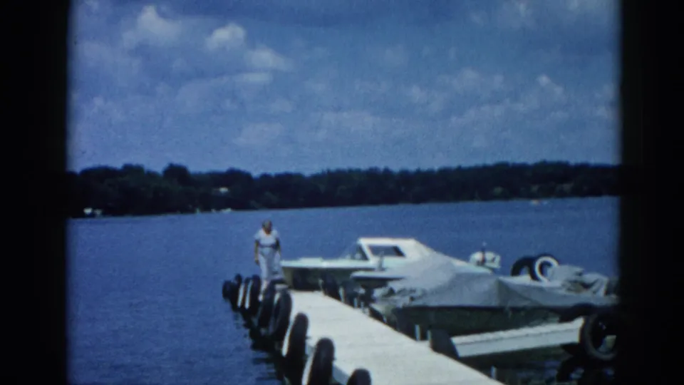 8mm frame showing a woman walking toward camera along a dock on an Illinois lake, circa 1962, with a white motorboat and covered boats moored alongside.