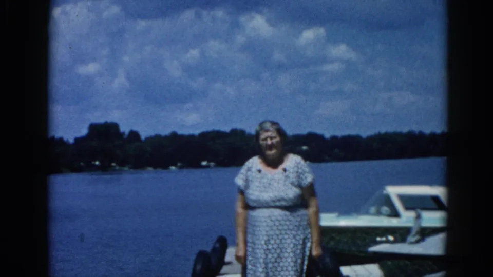 Close-up 8mm frame of a smiling older woman in a floral dress on a dock, Illinois lake circa 1962; white motorboat and tree-lined shore visible behind her.